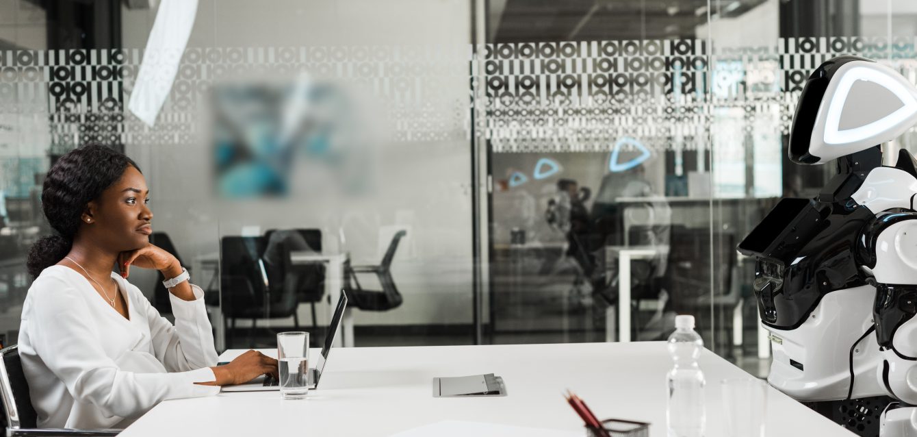 smiling african american businesswoman and robot sitting at desk in conference hall smiling african american businesswoman and robot sitting at desk in conference hall