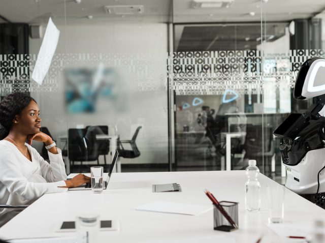 smiling african american businesswoman and robot sitting at desk in conference hall